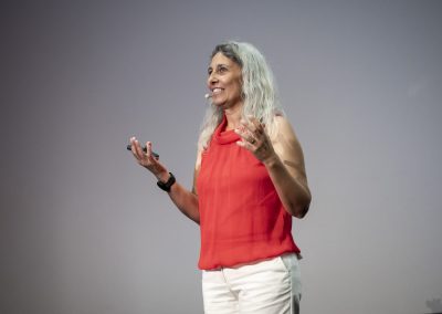 Speaker on stage, a woman with gray hair in an orange sleeveless top and white pants, wearing a headset mic and gesturing with her hands.
