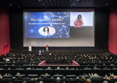 Großer Kinosaal mit einer Präsentation: Auf der linken Leinwand steht der Text ‚Was fühlst Du gerade?‘, rechts eine Moderatorin in rotem Oberteil vor Publikum.