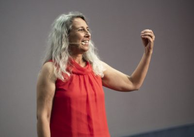 Smiling woman with long gray hair in a red sleeveless dress, speaking on stage with a headset mic.