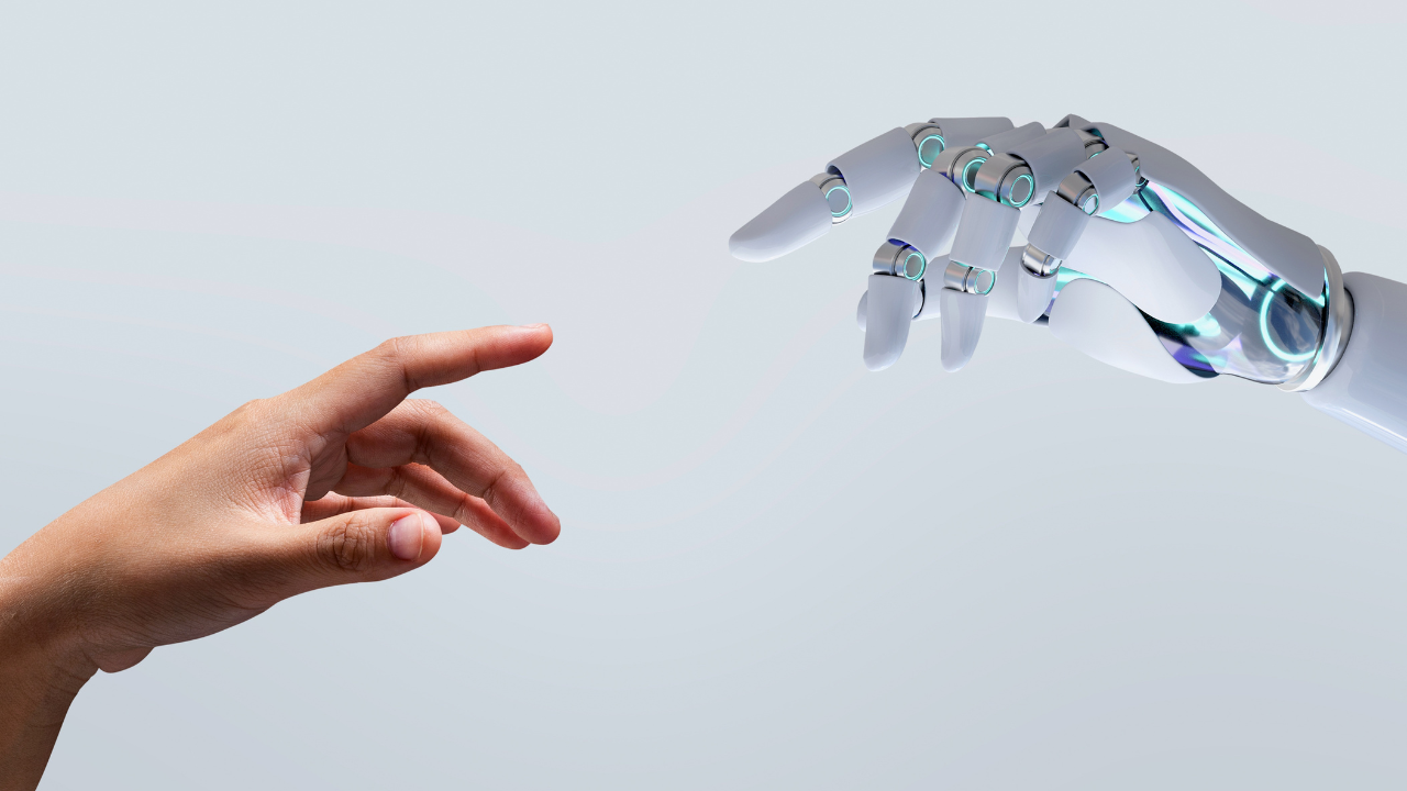 Human hand reaching toward a metallic robotic hand, symbolizing human–machine collaboration against a pale background.